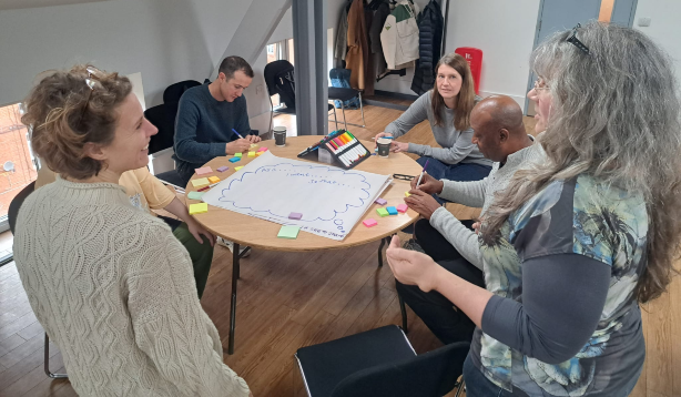 A small leadership team are gathered arounf a table working on an exercise with post it notes. One woman and Julia are standing and talking and smiling. the team is small but diverse.