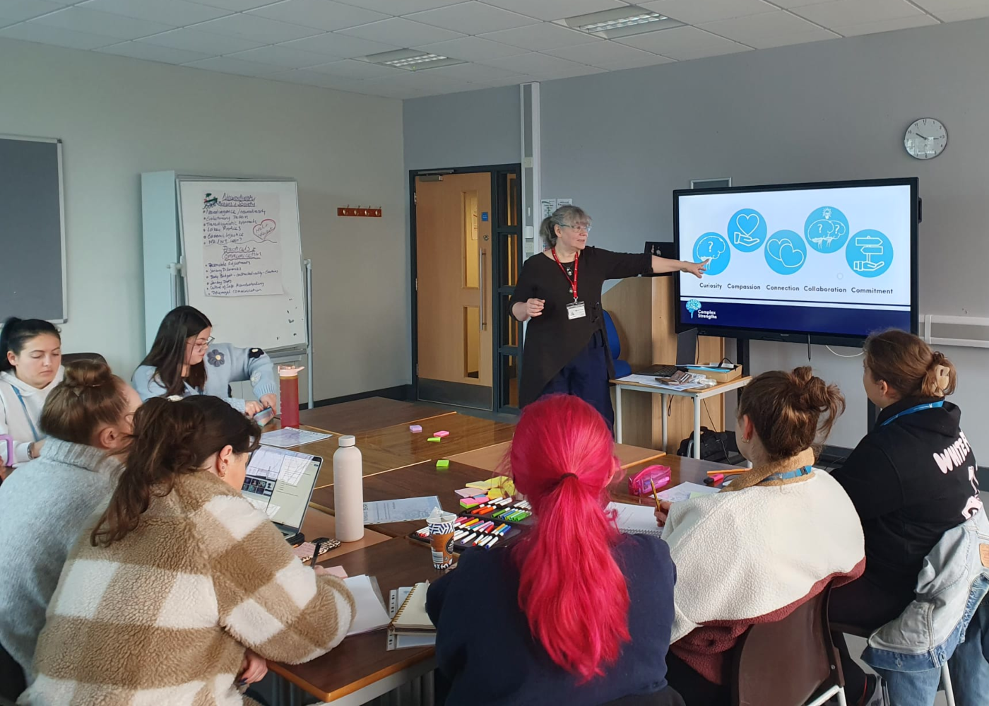 A small leadership team are gathered arounf a table working on an exercise with post it notes. One woman and Julia are standing and talking and smiling. the team is small but diverse.